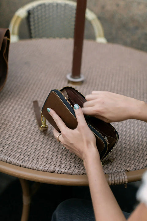 A person with light blue nail polish opens the Urban Southern Urban Zipper Wallet with card slots while seated at a round, woven outdoor table.
