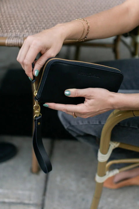 A person with mint green nail polish holds the Urban Southern Urban Zipper Wallet, a black leather wallet with credit card slots, while sitting on a woven chair.