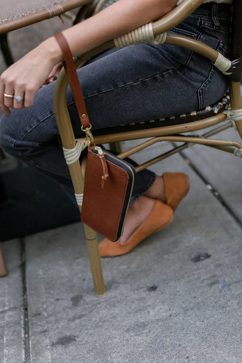 Seated outdoors on a woven chair, a person in black jeans and tan flats has an Urban Southern Urban Zipper Wallet—featuring an accordion design and visible card slots—hanging from the chair, with only their lower body and one arm shown.