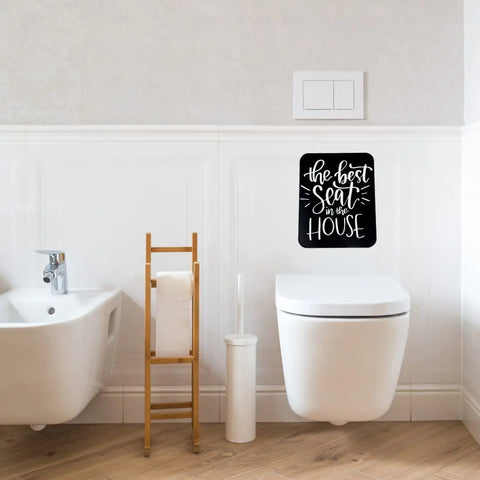 Modern bathroom with a white toilet, a Chatelet Manufacturing The Best Seat in the House black & white funny bathroom sign above it, plus a toilet paper holder, toilet brush, and bathtub on wooden flooring.