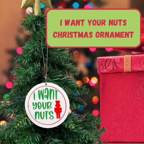 A Christmas tree displays the Chatelet Manufacturing Funny Gag Gift Christmas Ornament—perfect for Dirty Santa—reading “I want your nuts” in green and red, beside a pink-wrapped present and colorful festive holiday lights in the background.