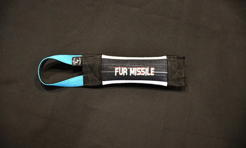 The Bulletproof Pet Products Inc Fur Missile Fire Hose Pocket Tug is a black and blue dog toy with a handle and FUR MISSILE in white and red letters on black, shown lying on a solid black surface.