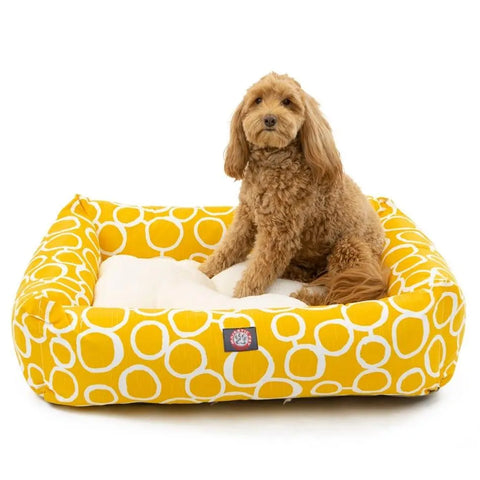 A small, curly-haired brown dog sits on the Majestic Pet Fusion Luxe Bagel Bed, which features white circular patterns and a soft white cushion, looking toward the camera.