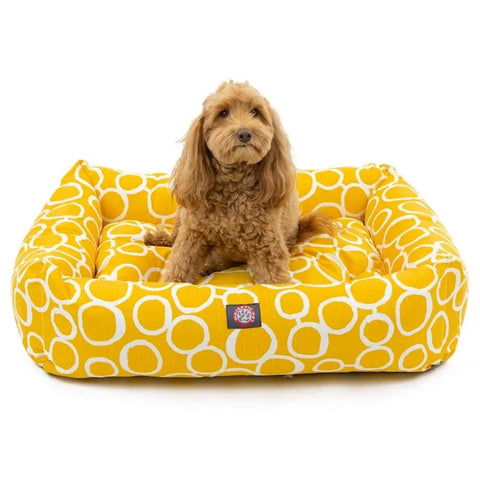 A small, fluffy brown dog sits on the Majestic Pet Fusion Luxe Bagel Bed, which is yellow with a white circular pattern and is machine washable.