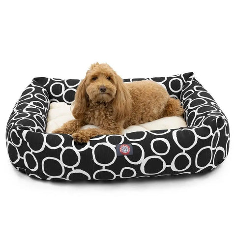 A small, fluffy brown dog is lying on the Majestic Pet Fusion Luxe Bagel Bed, featuring a luxurious black and white circular pattern, looking toward the camera.