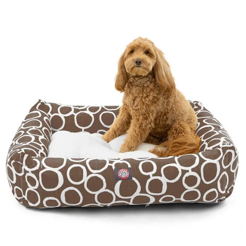 A fluffy brown dog sits in the Majestic Pet Fusion Luxe Bagel Bed, featuring white circular patterns and a soft white cushion. The luxurious bed is machine washable, and the dog looks toward the camera.
