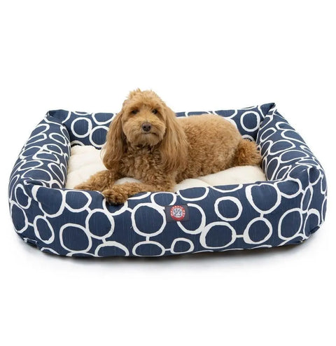 A small, curly-haired brown dog is lying on the Majestic Pet Fusion Luxe Bagel Bed, which features a cushioned center and a stylish navy blue and white pattern, looking toward the camera.