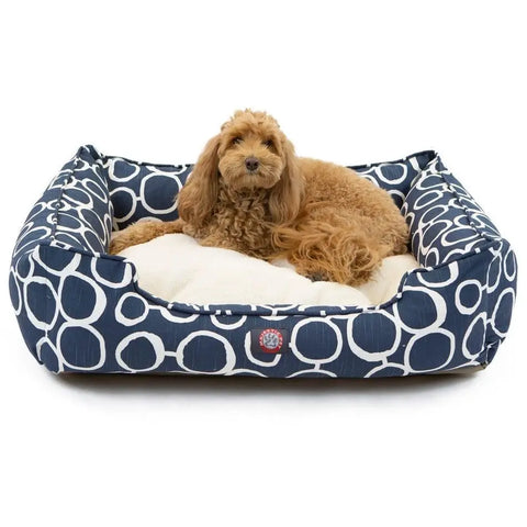 A fluffy, brown dog relaxes on a soft, cream cushion inside the Majestic Pet Fusion Contemporary Lounge Bed—blue with a white circle pattern, durable upholstery, and machine washable for easy cleaning—as it looks toward the camera.