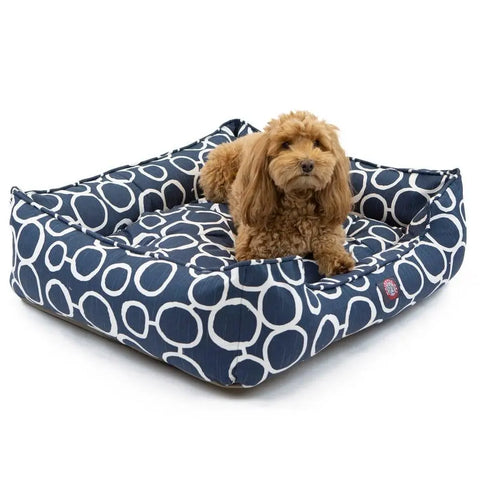A small, curly-haired brown dog is lying on the Majestic Pet Fusion Contemporary Lounge Bed, featuring durable upholstery, raised sides for coziness, and a machine washable design to keep it fresh and comfortable.