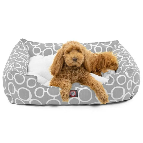 A small, curly-haired brown dog lounges on the Majestic Pet Fusion Contemporary Lounge Bed with white circular patterns, front paws on the edge, looking toward the camera.