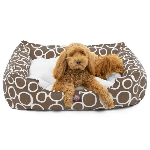 A small, curly-haired brown dog relaxes on the Majestic Pet Fusion Contemporary Lounge Bed, featuring a durable, machine-washable cover with a brown and white circular pattern and a soft white cushion for comfort.
