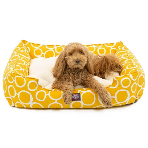 A small, curly-haired brown dog rests in the Majestic Pet Fusion Contemporary Lounge Bed, featuring a vibrant yellow color with white circle patterns. The bed is machine washable, soft, and comfortable as the dog looks directly at the camera.