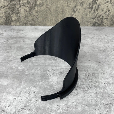A black Gallon Paint Pourer by Chatelet Manufacturing, designed to fit gallon-sized paint cans and prevent spills, with two prong-like ends, sits on a gray textured surface against a mottled wall.
