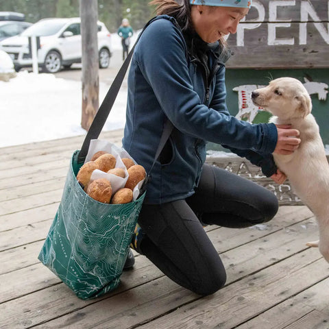 A person kneels on a wooden deck, smiling and petting a small light-colored puppy. They carry an eqpd GearBucket - Methow Map watertight tote bag with bread rolls. Cars and trees can be seen in the background.