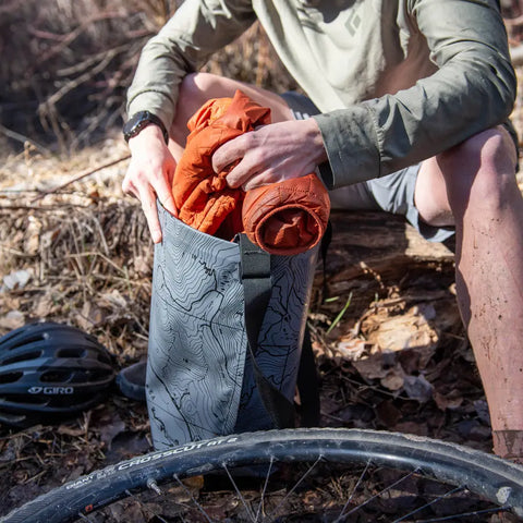 A person sitting on a log with an eqpd GearBucket - Methow Map, a watertight tote bag, full of clothes.
