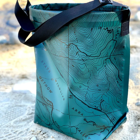 The eqpd GearBucket - Methow Map is a green tote bag with black straps, a structured rim for durability, and a detailed topographic map design featuring contour lines, labels, and geographic features.