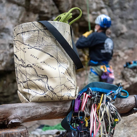 A climber with harness and helmet stands in the background, while the eqpd GearBucket - Methow Map watertight tote bag and a harness with carabiners and ropes hang from a wooden log in the foreground.