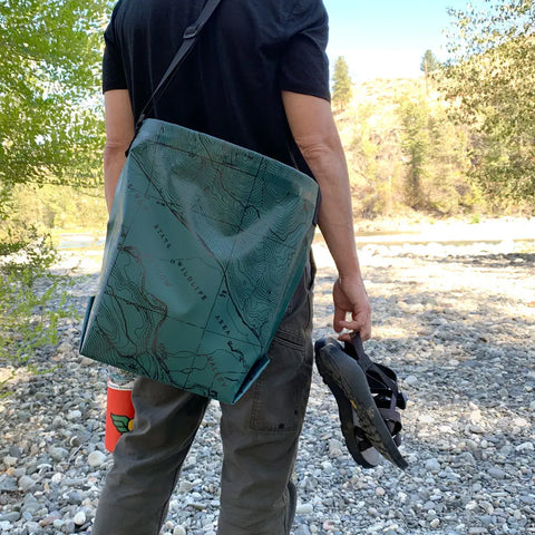 Outdoors on rocky ground, a person holds sandals and a cup with the eqpd GearBucket - Methow Map (featuring a watertight design, structured rim, and removable felt base) at their side; trees and a sunlit hillside appear in the background.