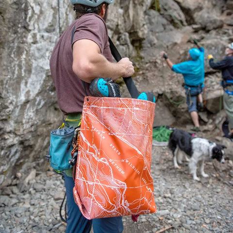 A climber carries the eqpd GearBucket - Methow Map, a watertight tote with an orange topographic map design, packed with a helmet and shoes near rocks where other climbers and a dog are present.