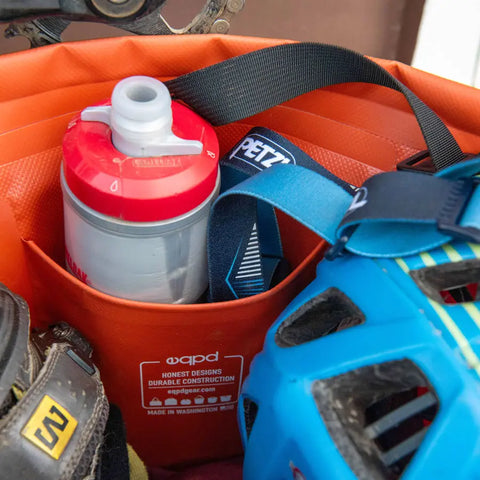 A close-up of the eqpd GearBucket - Methow Map shows its structured rim keeping contents—red and white water bottle, blue bike helmet, black strap—organized. A label is visible on the pocket of this watertight tote bag.