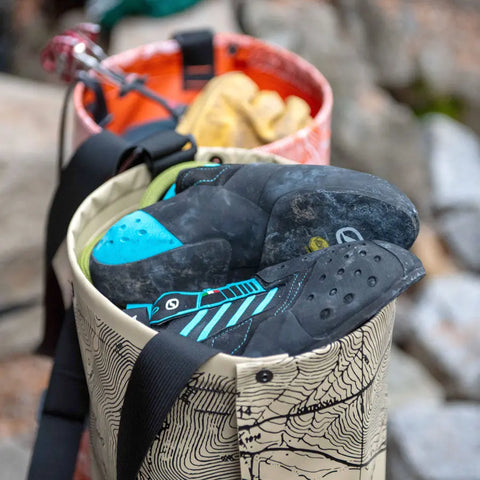 A close-up of two eqpd GearBucket - Methow Map bags; the front holds black and turquoise climbing shoes, the back stores climbing gear and yellow chalk, all outdoors on rocky terrain.