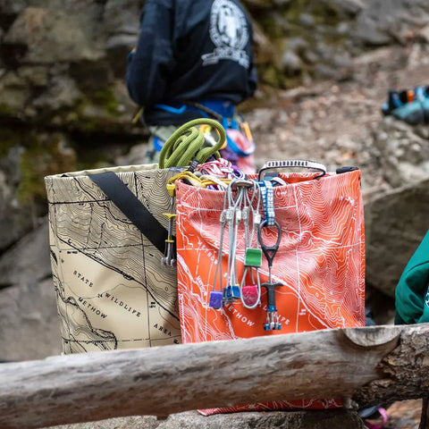 Two eqpd GearBucket - Methow Map bags with structured rims and topographic designs sit outdoors, holding climbing ropes and hardware. A harnessed person stands nearby, facing away.