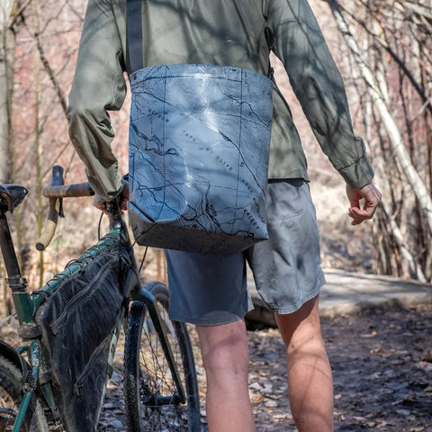 A person in a green jacket and gray shorts stands on a wooded trail with a bike, carrying the eqpd GearBucket - Methow Map, a watertight tote bag featuring a topographic map design and structured rim for shape and support.