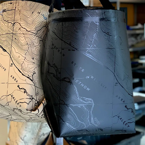 Two GearBucket - Methow Map totes by eqpd hang side by side, each with a unique topographic map design. The dark grey Methow tote stands out, and both include a removable felt base for extra support.