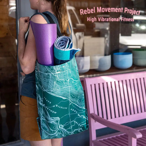 A person with an eqpd GearBucket - Methow Map tote featuring a green topographic print and structured rim stands near a Rebel Movement Project High Vibrational Fitness window, carrying a purple yoga mat and blue towel by a pink bench.