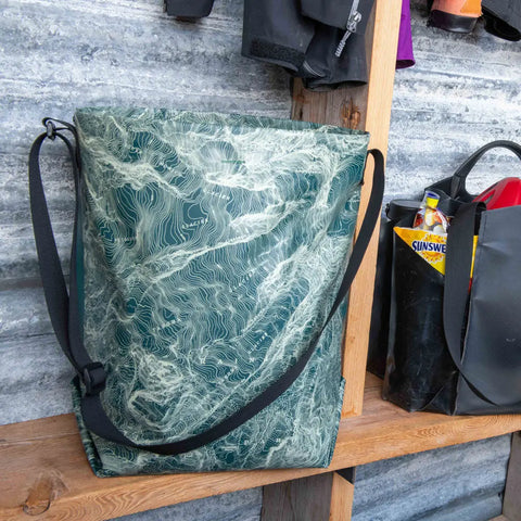 An eqpd GearBucket - Mount Rainier, featuring a green watertight design with a white abstract wavy pattern, hangs on a wooden shelf beside a black bag with various items, set against a corrugated metal wall.