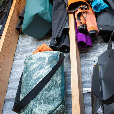 Colorful eqpd GearBucket - Mount Rainier bags and jackets are displayed on wooden shelves against a corrugated metal wall. Some bags have removable felt bases; others feature watertight tote designs with straps and patterns.