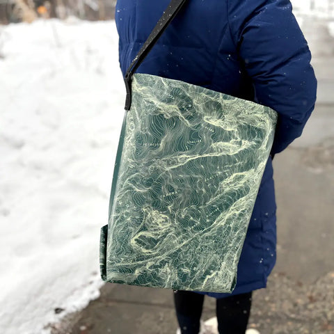 A person in a blue coat stands on a snowy sidewalk, carrying the eqpd GearBucket - Mount Rainier, a large green and white watertight tote featuring an abstract topographic map design.
