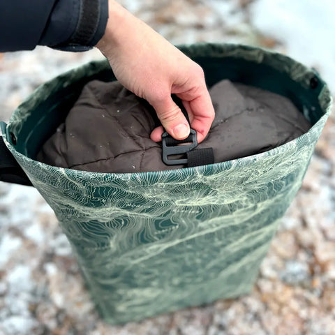 A hand clasps the buckle of an eqpd GearBucket - Mount Rainier, a green patterned watertight tote, filled with a brown quilted item and set outdoors on a snowy, rocky surface.