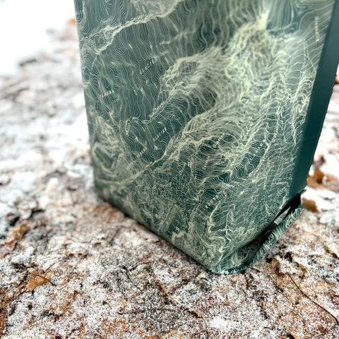 A close-up of the eqpd GearBucket - Mount Rainier, a structured green storage bin with a wavy white line pattern, resting on snow-dusted ground among scattered brown leaves.