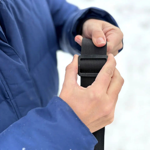A person in a blue jacket uses both hands to adjust the black strap with a plastic buckle on the eqpd GearBucket - Mount Rainier watertight tote bag; the blurred white background gives a snowy, outdoor feel.