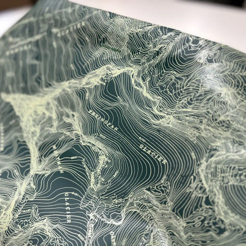 A close-up of a topographic map with detailed contour lines and labeled features evokes the textured surface of the eqpd GearBucket - Mount Rainier, creating a striking three-dimensional effect against a dark background.