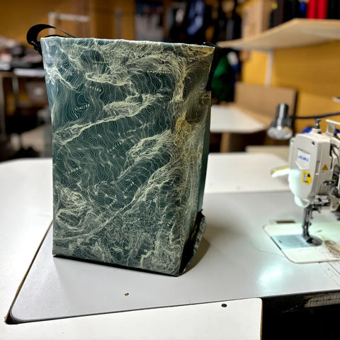 The eqpd GearBucket - Mount Rainier, a green and beige fabric bag featuring a topographic map design and removable felt base, is displayed on a sewing table beside a white sewing machine in a workshop setting.