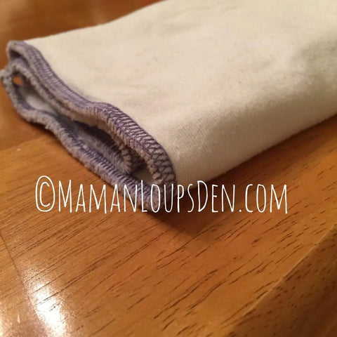 A Geffenbaby Jersey Cloth Diaper Prefold (60% hemp, 40% organic cotton) with purple stitching lies folded on a wooden surface. Text overlay: “@MamanLoupsDen.com.”.