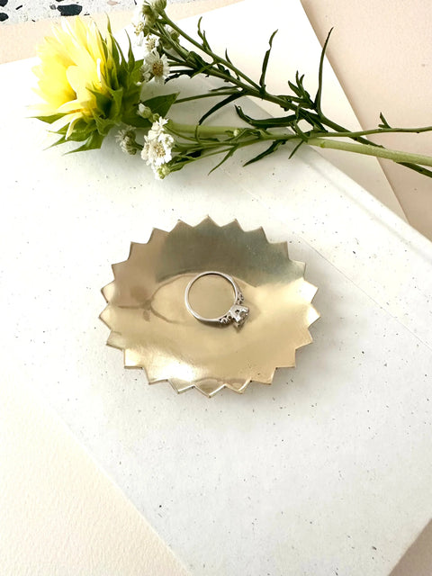 A silver ring with a single gemstone rests on the Sarah Cecelia BRASS POINTE JEWELRY DISH, a handmade metallic star-shaped piece. The dish sits on a white surface with a yellow flower and delicate white blossoms nearby.