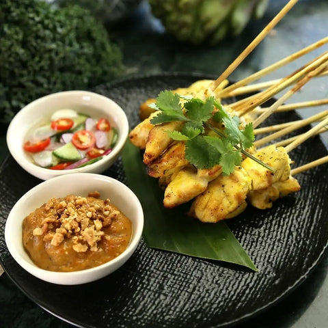 Grilled chicken skewers seasoned with Gustus Vitaes Global Foodie Collection - 4 Tins, garnished with cilantro, served on a black plate with peanut sauce topped with crushed peanuts and cucumber-chili relish.