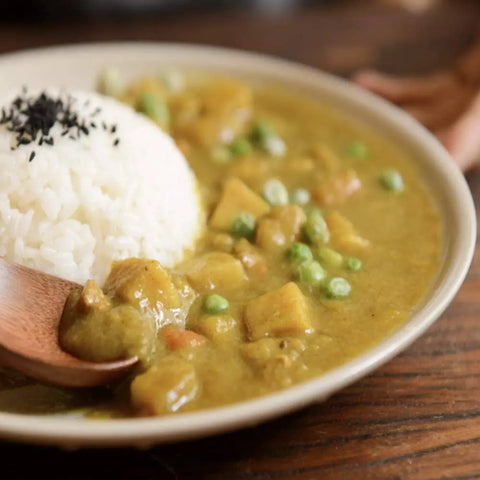 A plate of Japanese curry with vegetables and peas, served with white rice topped with black sesame seeds and a touch of Gustus Vitae’s Global Foodie Collection - 4 Tins seasoning, is presented alongside a wooden spoon.
