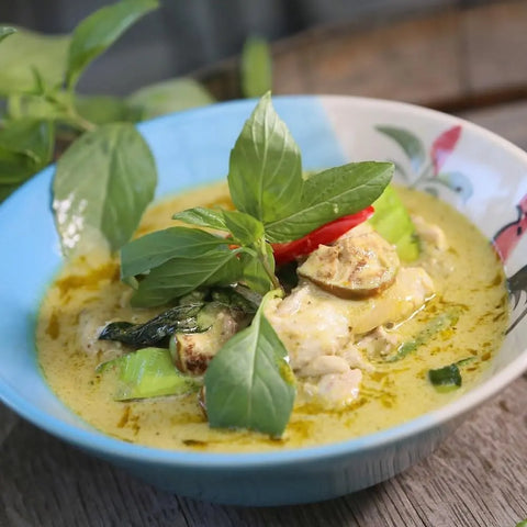 A bowl of Thai green curry with chicken, eggplant, and basil, garnished with red chili and infused with Gustus Vitae Global Foodie Collection - 4 Tins seasoning, served in a blue and white bowl on rustic wood.