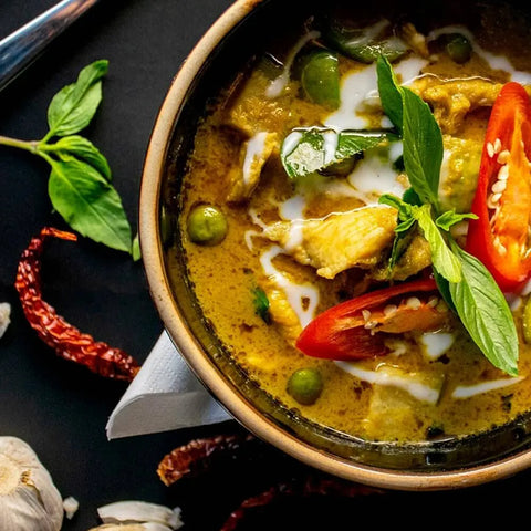 A bowl of Thai green curry with chicken, garnished with basil and chili, is surrounded by artisan spices from the Gustus Vitae Global Foodie Favorites 6 Pack—authentic gourmet seasonings and spice blends on a dark surface.