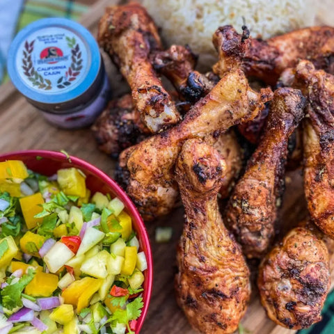 Grilled chicken drumsticks on a wooden board, paired with mango salad in a red bowl and white rice, are seasoned with Gustus Vitae’s Global Foodie Favorites 6 Pack—authentic gourmet spice blends from around the world.