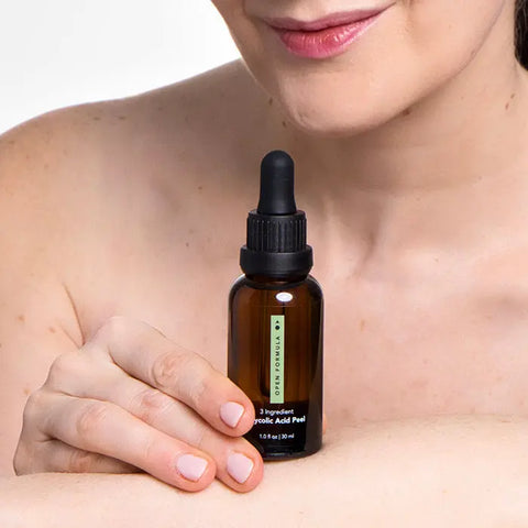 A woman with bare shoulders smiles gently while holding a brown dropper bottle of Open Formula Glycolic Acid Peel against her arm, showcasing this exfoliating solution for radiant, glowing skin.