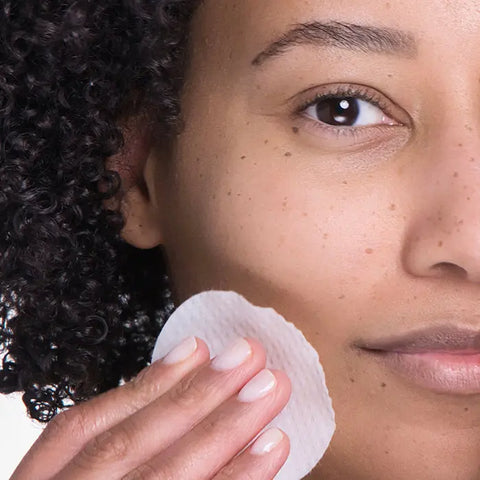 Close-up of a person with curly hair gently wiping their cheek with a round cotton pad, applying Open Formula Glycolic Acid Peel to exfoliate dead skin cells and reveal clear, smooth skin.