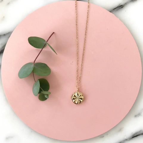 The GOLD HACIA PENDANT by Sarah Cecelia, a dainty gold necklace with a starburst design, is displayed on a round pale pink surface with eucalyptus, set against a white marble background to highlight its handcrafted elegance.