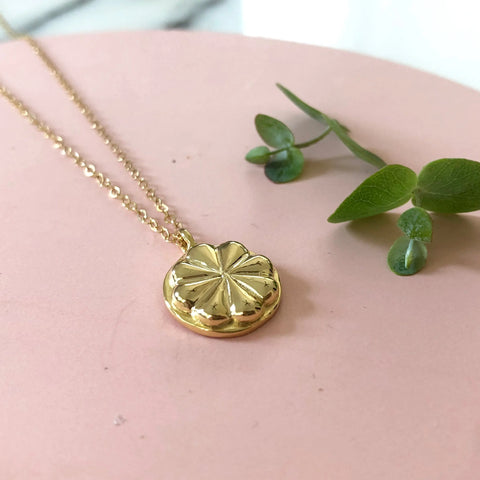 The GOLD HACIA PENDANT by Sarah Cecelia—a dainty gold necklace with a textured flower design—rests on a pink surface beside a green plant sprig, showcasing the unique charm of handcrafted jewelry.