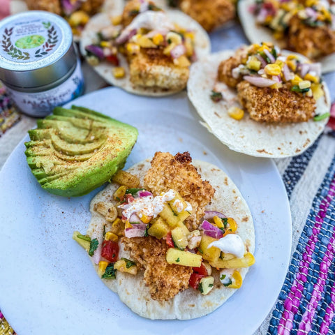 A plate holds a sliced avocado and an open-faced taco with breaded fish, corn salsa, sauce, and Gustus Vitae gourmet sea salt from the Ultimate Artisanal Seasoning and Gourmet Sea Salt Collection - 20 Tins, with extra tacos and a tin in the background.