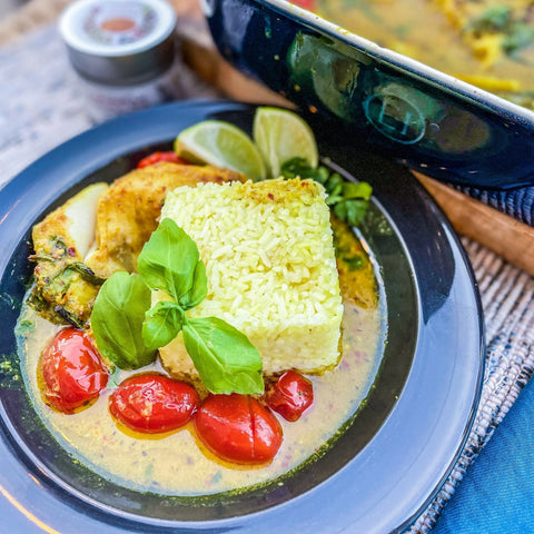 A plate with square yellow rice garnished with basil and finished with Gustus Vitaes Ultimate Artisanal Seasoning and Gourmet Sea Salt Collection, surrounded by roasted cherry tomatoes, creamy chicken, lime wedges, and a blue casserole dish nearby.